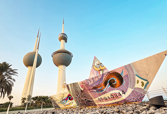 Kuwait Towers are seen at sunset with an artistic sculpture shaped like a paper boat in the foreground.